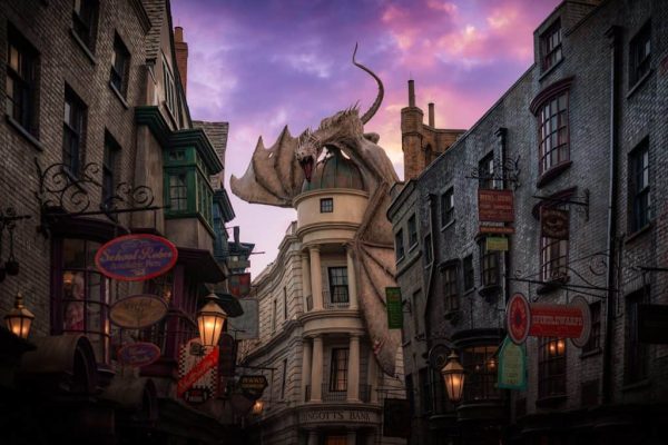 Walking into Diagon Alley. Striaght ahead is Gringotts bank with the dragon on the top. Lining either side of the photo are shops. There is a beautiful sunset in the background. 